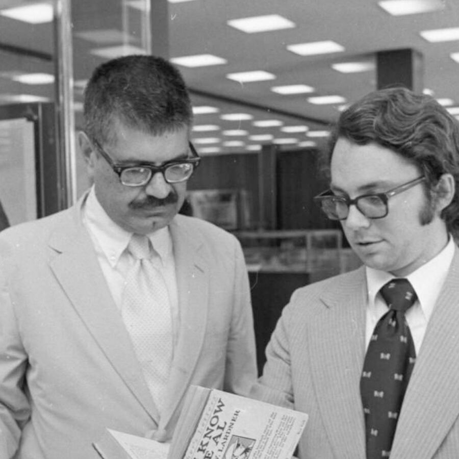 Black-and-white photograph from the archive of two men in suits reading a printed sheet.